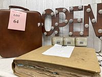 Photo showing a brown vintage bar suitcase, metal 'OPEN' sign with light bulbs, and large 1950s scrapbook with worn pages on a white surface.