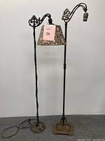 Pair of vintage cast iron floor lamps shown side by side against a plain wall and grey carpet. The left lamp includes a floral fabric lampshade, the right lamp is missing the lampshade. Both lamps stand approximately 5 feet tall, with ornate cast iron bases and stands.