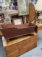 Side view of the primitive wooden cradle sitting on a wooden chest, showing its rectangular body and dark wood finish.