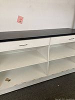 Front view of the retail counter showing black countertop, white drawers with metal handles, and open shelving beneath. Shows wear and chipping on shelving base.