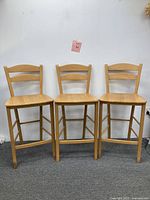 Full view of all three wooden ladder-back counter height bar stools side by side against a white wall with visible floor.