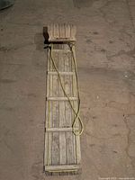 Full length top down view of the wooden toboggan on a wooden floor showing the rope handle and slatted wood construction.