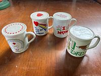 Front view of 4 white ceramic mugs with lids on a wooden surface, showing colorful printed designs related to love, family, and positivity.