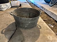 The galvanized metal pot shown from a slightly side angle highlighting one handle and the overall shape, sitting on a concrete floor in a warehouse-like area.