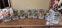 Nine clear glass Champion beer mugs displayed in a row on a wooden table, showing logos and slogans clearly.