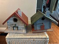 Front view of two handmade wooden bird houses on a wicker surface, showing bird entry holes and perch on the white/red house.