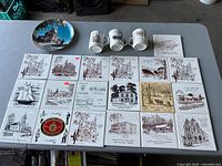 Overview of all items laid out on table, including 21 trivets, 3 mugs, 1 decorative plate, and 1 ceramic tile