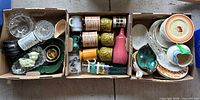Three cardboard boxes containing various glassware, pottery, and ceramic items including mugs, plates, bowls, and decorative pieces. Items have varied colors and patterns, some visible damage and wear.