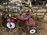 Side view of red Radio Flyer tricycle showing chrome handlebars with surface rust and red plastic seat and body with dirt.