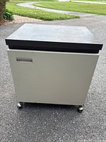 Front view of the metal rolling office cabinet showing grey body, black top, and caster wheels.