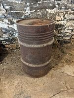 Full side view of the rusty metal barrel showing ridges and surface corrosion against a stone wall background.