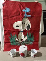 Snoopy wall hanging held up to show full design and size, with cookie jar and mugs in front.
