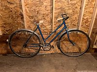 Full side view of blue vintage Supercycle bicycle leaning against plywood wall, showing frame, wheels, handlebars, and saddle.