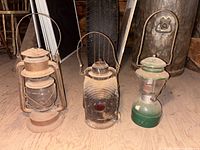 Three old lanterns on wooden floor: older metal and glass lamp with 'Made in Canada' stamp and Wright logo on base, black signal lantern missing lid with red glass beacon, and vintage Coleman lantern with green base.