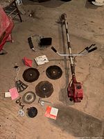 Overview photo showing brush cutter engine with handlebars, multiple circular blades, plastic guard, small parts, and manual on floor.