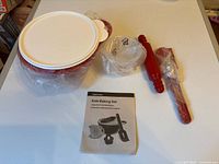 Full view of all items including mixing bowl with lid, measuring cup, rolling pin, spatula, flower-shaped item, and instruction booklet.