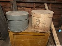 Two antique wooden cheese boxes displayed side by side on a rustic wooden chest, showing front and side views with visible handwritten markings on one box.
