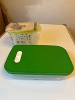 Photo of two plastic food storage containers on a white surface, one with a green lid and the other still in plastic wrap with a yellow-green lid.
