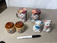 Photo showing two pairs of salt and pepper shakers: one floral patterned in metal holders and one of white ceramic cats with bows; marker for size reference