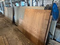 Multiple large sheets of plywood and paneling stacked against a wall in a basement or garage space, showing overall size and condition.