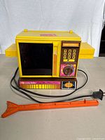 Front view of yellow vintage Betty Crocker Easy Bake Four Mini-Wave Oven with side trays, keypad, dial timer, and clear door window. Cable and orange pushing stick in front.