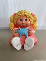 Front view of vintage Cabbage Patch Kids blonde hair, blue eye girl plastic coin bank holding a blue pig, showing wear and condition.
