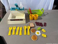 Full set showing plastic sink with blue faucet, soap dispenser missing cap, green dish rack with red utensils, two yellow cups, and various food pieces including utensils and foods.