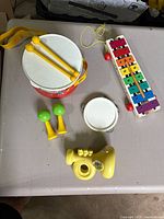 Top-down view showing the Fisher Price red drum with yellow strap, yellow drumsticks, white tambourine, colorful xylophone with red mallets, green and yellow maracas, and yellow plastic trumpet with Smurf stickers missing.