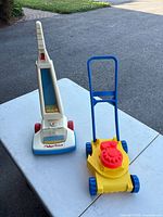Both vintage toy vacuum and plastic toy lawn mower displayed on a white table outdoors, showing overall condition.