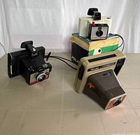 Three instant cameras displayed on white surface: Polaroid Colorpack 80, Polaroid Swinger Model 20, Kodak Pleaser Instant Camera arranged with box in background