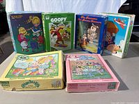 Six vintage puzzle boxes displayed standing or lying flat showing different characters: Rainbow Brite, Goofy, The Raccoons, Porky Pig, Cabbage Patch Kids, Rose-Pedal Place.