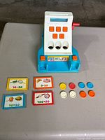Front view of the vintage Chicco plastic toy cash register with orange buttons, blue base, and red side crank. Coins and cards are displayed in front.