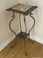Full view of antique brass and marble two-tiered display stand against a wall with wood floor, showing side angle view.