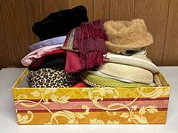 Box filled with various women's hats and scarves including fur hats, leopard print scarf, straw hats, and colorful scarves.