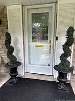 Frontal view of both black cast iron urn planters with faux spiral topiary shrubs positioned in front of a white door