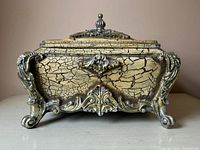 Front view of the decorative ornate wooden box, showing the crackled paint finish and intricate metallic accents on the front and feet.