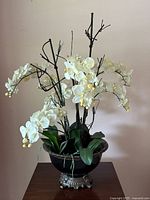 Front view of the full faux orchid flower arrangement in the black glazed jardiniere with ornate metal base on a wooden table against a pink wall.