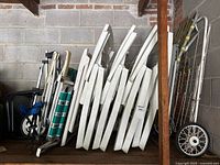 Side view showing nine white plastic lawn chairs stacked, white folding golf cart and grocery buggy with wheels on the right side, and some green crates on the left.