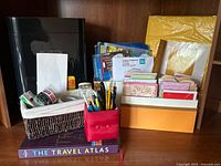 Photo of paper shredder, greeting cards, stationery, tape rolls in basket, pencils in fabric holder, and The Travel Atlas book on a shelf
