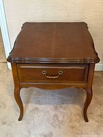 Front view of one walnut side table showing scalloped top, single drawer with metal handle, and cabriole legs.