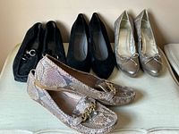 Four pairs of Stuart Weitzman women's shoes arranged on a pale cream table: taupe snakeskin loafers with gold chain, two pairs of black suede shoes (one loafers with silver hardware and one pumps), and a metallic silver pump with bow detail.