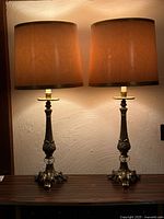 Pair of vintage brass table lamps with lit brownish-beige fabric shades on dark wood surface against textured wall.