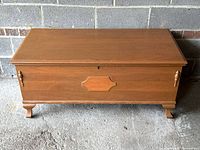 Front view of the closed cedar lined wooden chest showing the mild scratches and warm wood finish, raised on decorative legs with side handles.