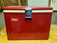 Front view of vintage Coleman red metal cooler with white trim and metal latch on a wooden surface.