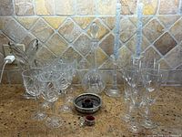 Wide shot of all glassware items on counter including decanter, wine glasses, champagne flutes, candlestick holders, vase and silver plated coaster.