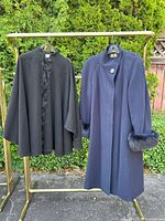 Black wool poncho with fox fur trim and navy wool coat with fox fur cuffs on gold clothing rack outdoors