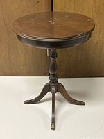 Front view of vintage round wood end table showing the round tabletop, turned center pedestal, and three curved legs with brass feet caps. Visible surface marks and stains on wood.