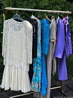 Full view of rack with seven 1980s women's beaded dresses and purple evening wear set hung outdoors on a gold clothing rack.