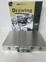 Front view of metallic silver sketching art case with ArtSchool Drawing Book in background