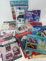 Image showing a variety of vintage crafts magazines including Mary Maxim catalogs, festive rub-on transfers, cross stitch pattern books, and packaged party game kits with EK tools embellishments.